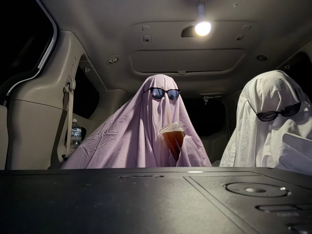 Two sheet ghosts in sunglasses riding in the back of a van at night, drinks in hand