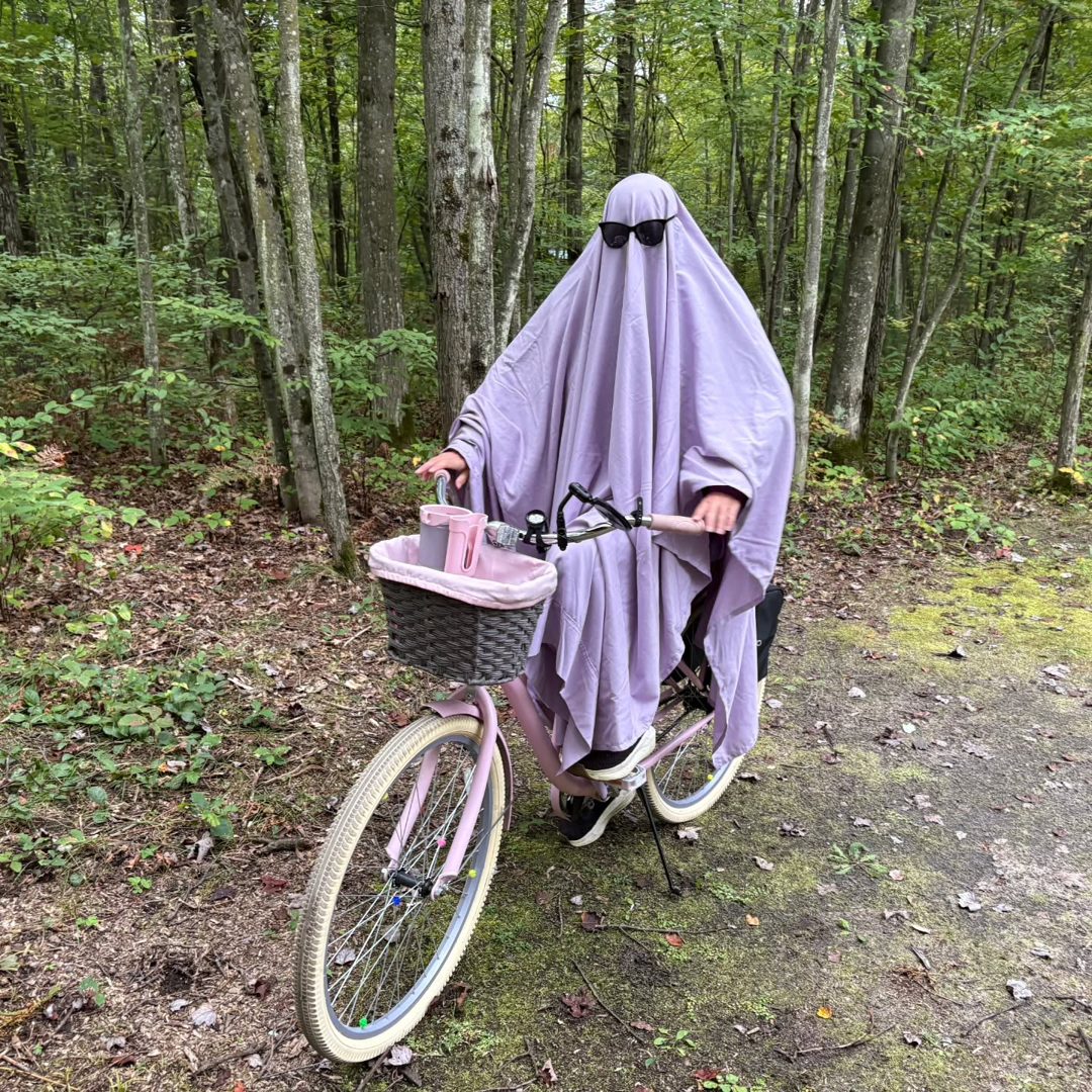 A lavender sheet ghost in sunglasses riding a pink bicycle with a basket down a forest trail